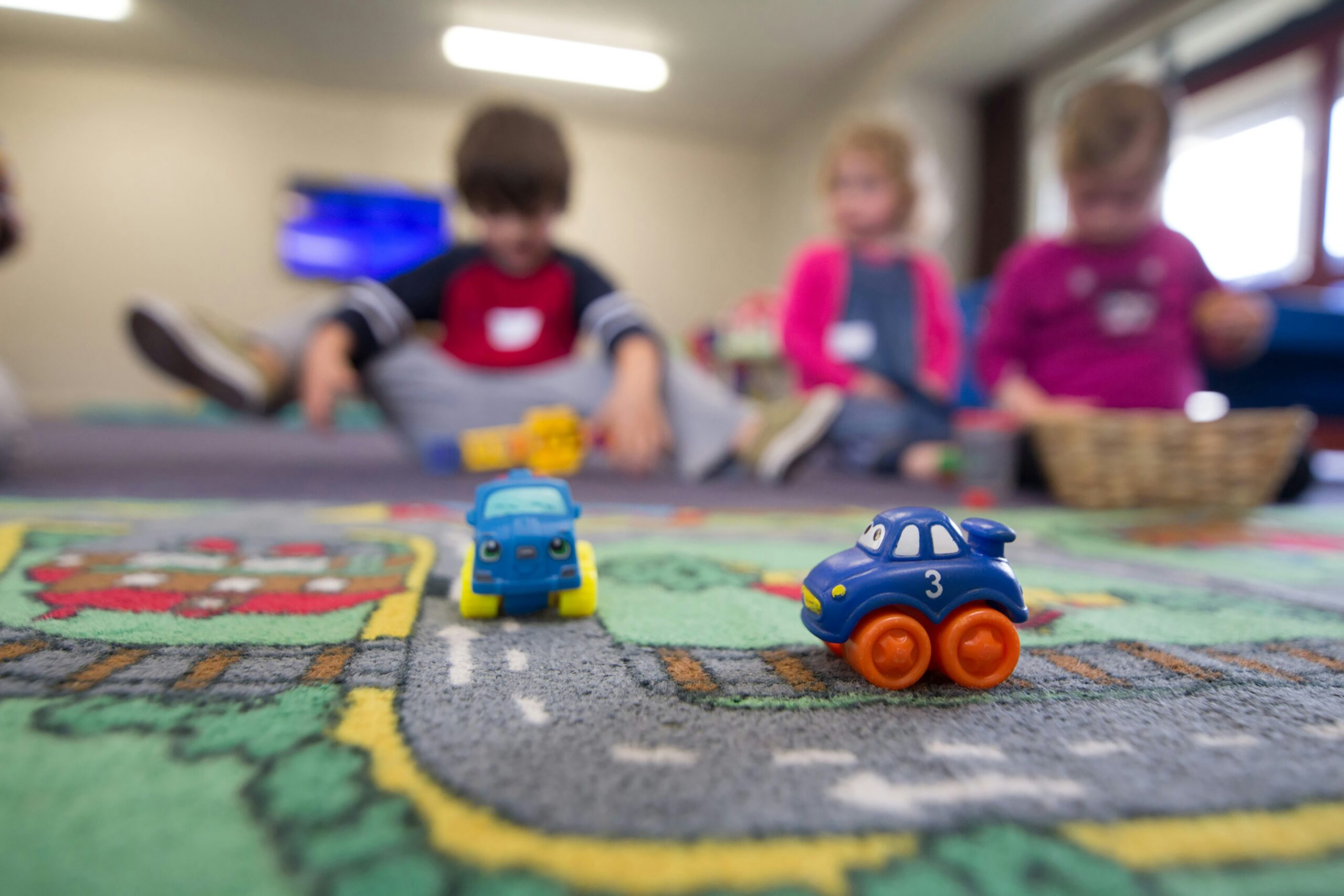 Image of pre-school children in the background, and toy cars in the foreground on a rug with a road pattern